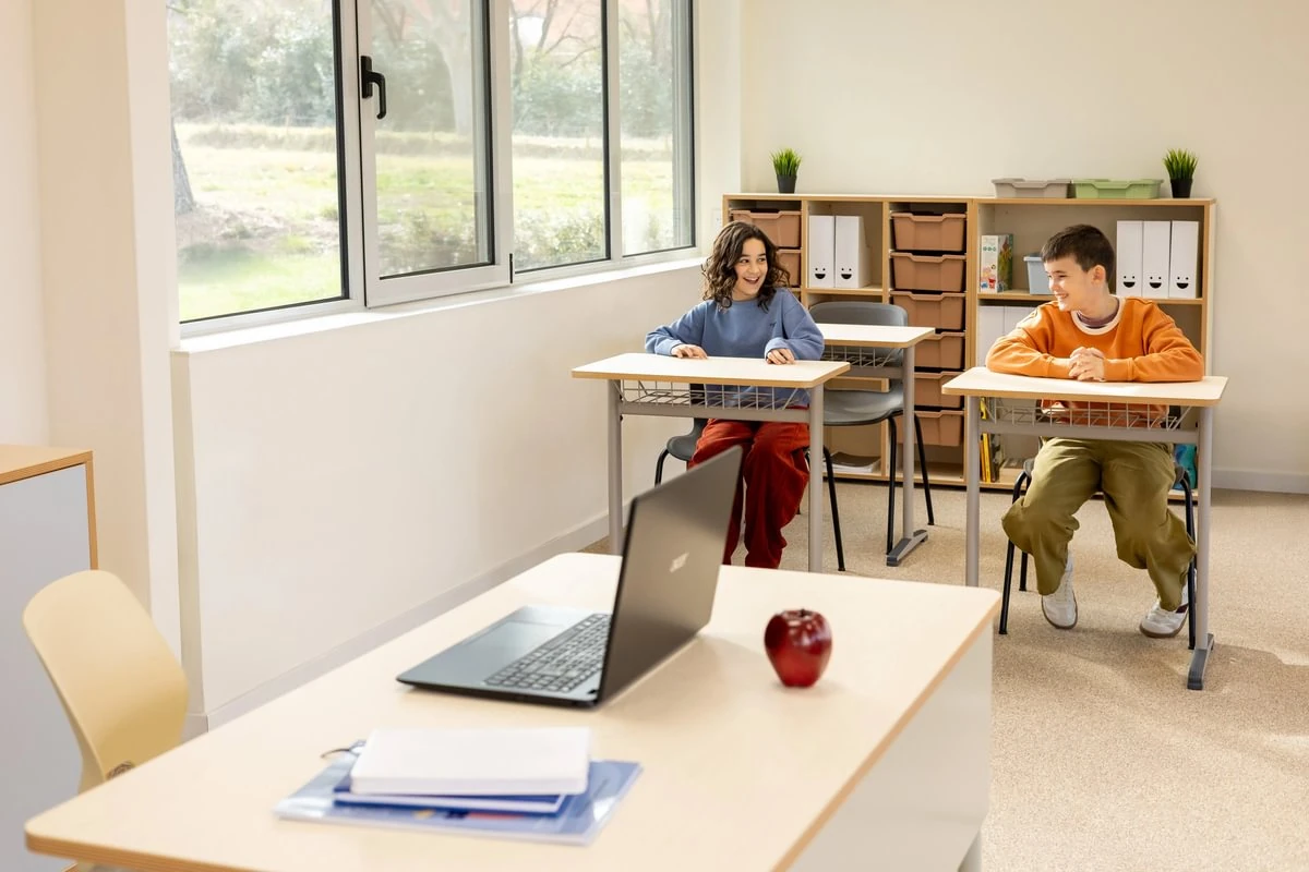 Schultische mit weißen Tischplatten und Drahtkörben in einem hellen Klassenzimmer