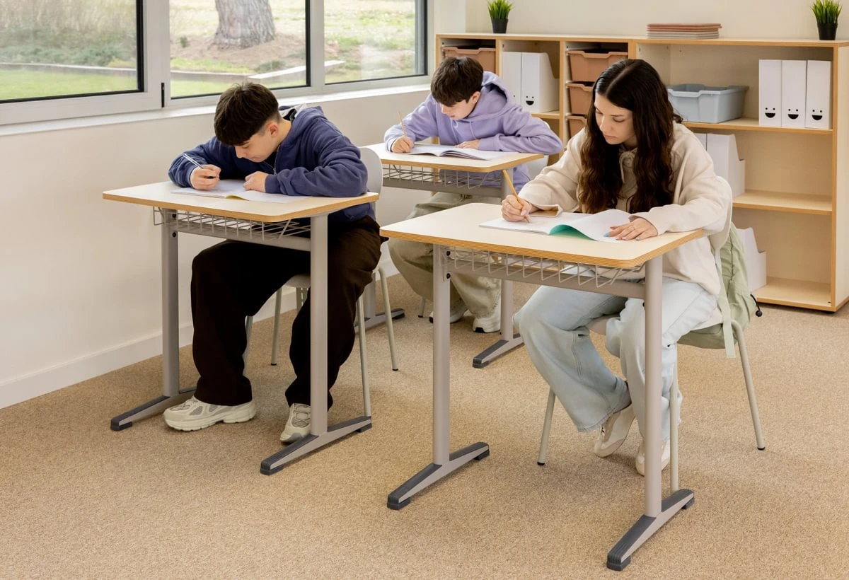Drei Schultische mit weißen Tischplatten und Drahtkörben während einer Schreibaufgabe im Klassenraum